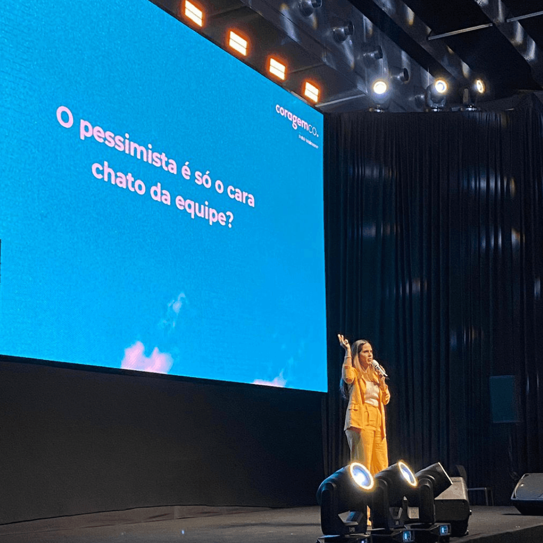 Speaker in orange suit addressing audience on stage with large screen showing text in Portuguese.